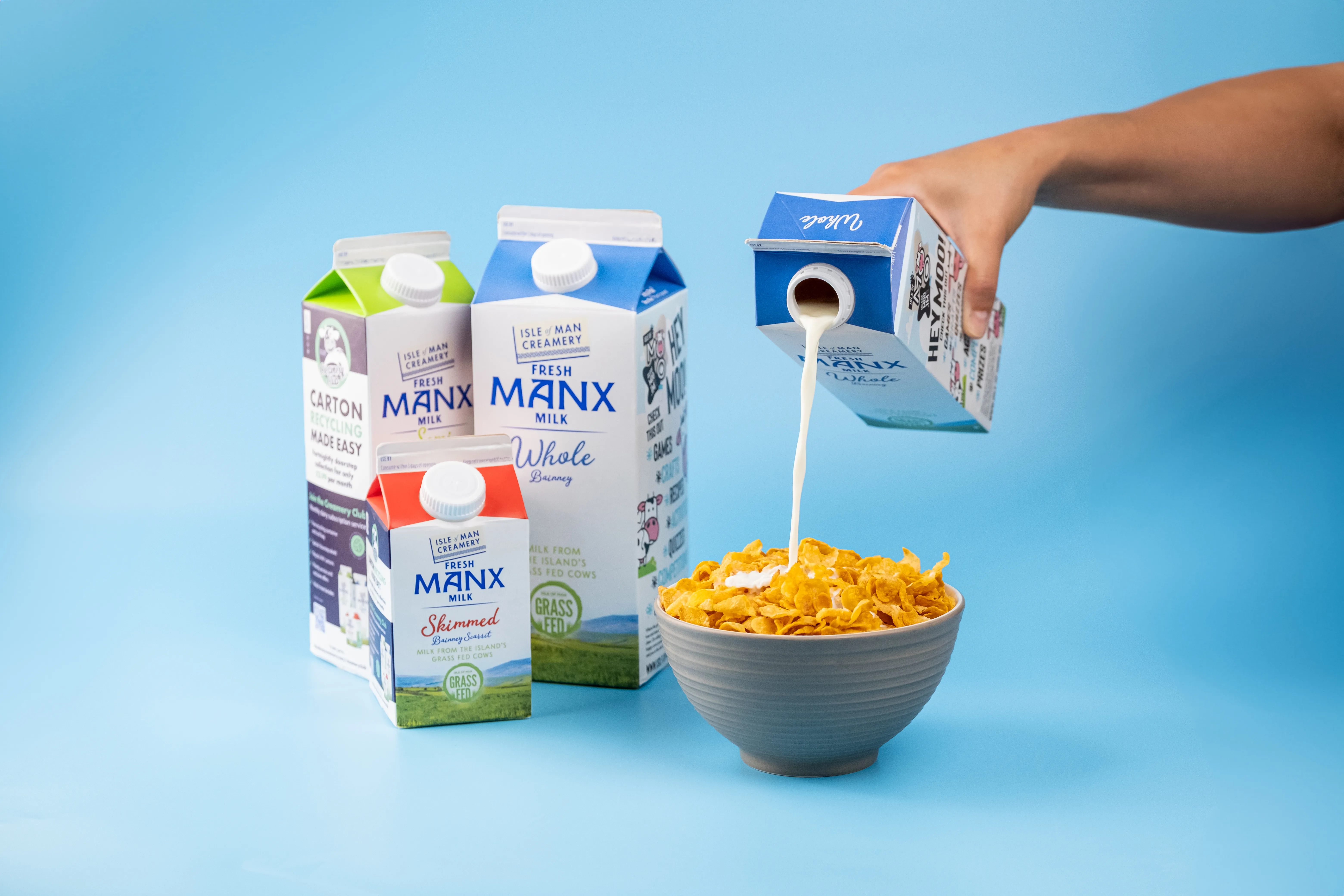 A hand pouring Manx Creamery whole milk from a carton over a bowl of cornflakes, surrounded by a range of Manx milk cartons including skimmed and semi-skimmed varieties, on a blue background